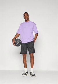 Lilla bomulls-t-skjorte med brodert logo, svarte mesh-shorts og svart-hvite sneakers med hvite sokker. Holder en basketball.