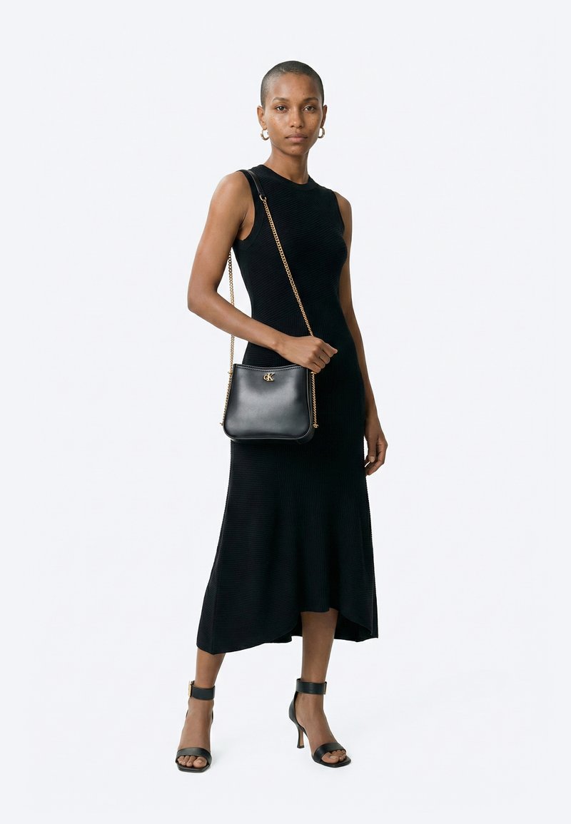 Femme en robe midi noire sans manches et sandales noires à talons hauts tenant un sac à bandoulière en cuir noir avec une chaîne dorée.