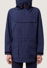 Marineblauwe waterdichte jas met een grote voorzak, verstelbare capuchon en elastische manchet. Voorzien van een drukknoopsluiting en merkpatch.