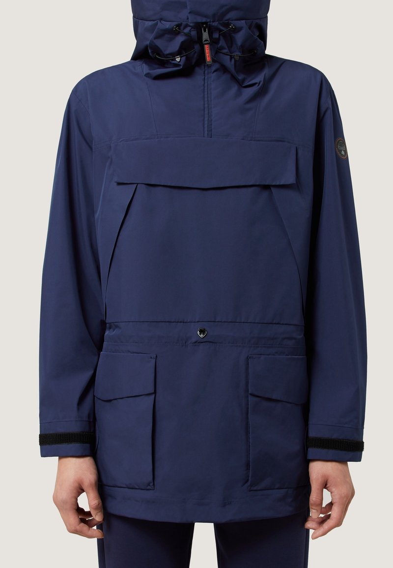 Marineblauwe waterdichte jas met een grote voorzak, verstelbare capuchon en elastische manchet. Voorzien van een drukknoopsluiting en merkpatch.