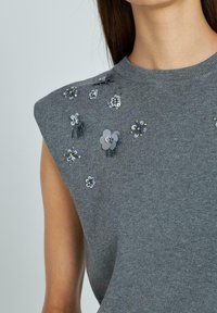Suéter gris sin mangas con textura, adornado con elementos florales en tonos variados de gris y plata. Los detalles incluyen lentejuelas y cuentas.