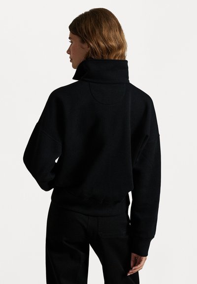 Svart hettegenser med høy krage og senkede skuldre, med en avslappet passform og ribbestrikket kant. Stoffet virker mykt og koselig.