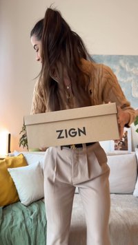 Mladá žena s dlouhými vlasy drží krabici od bot ZIGN, stojí v útulném obývacím pokoji s polštáři a rostlinami v pozadí.