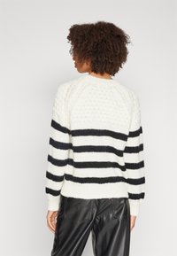 Suéter en crema con rayas horizontales negras, textura de punto, puños acanalados y detalles en la parte posterior. Combinado con pantalones de cuero negros.