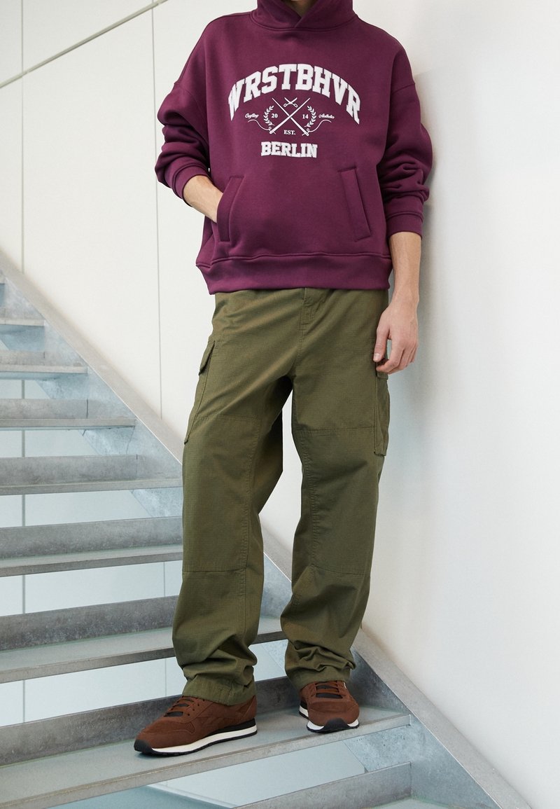 Bordeaux hoodie met witte grafische tekst en kangoeroezak; olijfgroene cargobroek; bruine sneakers met witte accenten, staande op een trap.
