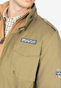 La chaqueta verde oliva presenta un cierre con cremallera, epaulettes en los hombros y dos bolsillos en el pecho. Fabricado en un tejido texturizado con un logo bordado de "HARPER".