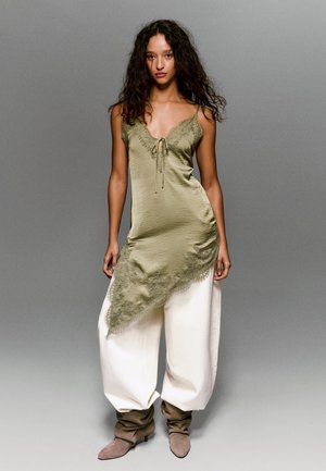 Vrouw met krullend haar die een asymmetrische slipjurk met kanten rand in olijfgroen draagt, gecombineerd met witte wijde broekspijpen en bruine slouchy laarzen.