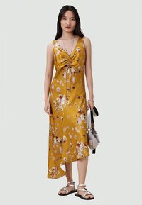 Gelbes Blumenkleid mit V-Ausschnitt, gerafftem Frontdetail und asymmetrischem Saum. Hergestellt aus Satin, kombiniert mit silbernen Sandalen und einer Tasche.