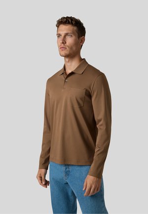 Mann trägt ein braunes Langarm-Poloshirt mit Brusttasche und blaue Jeans, steht vor einem schlichten grauen Hintergrund.