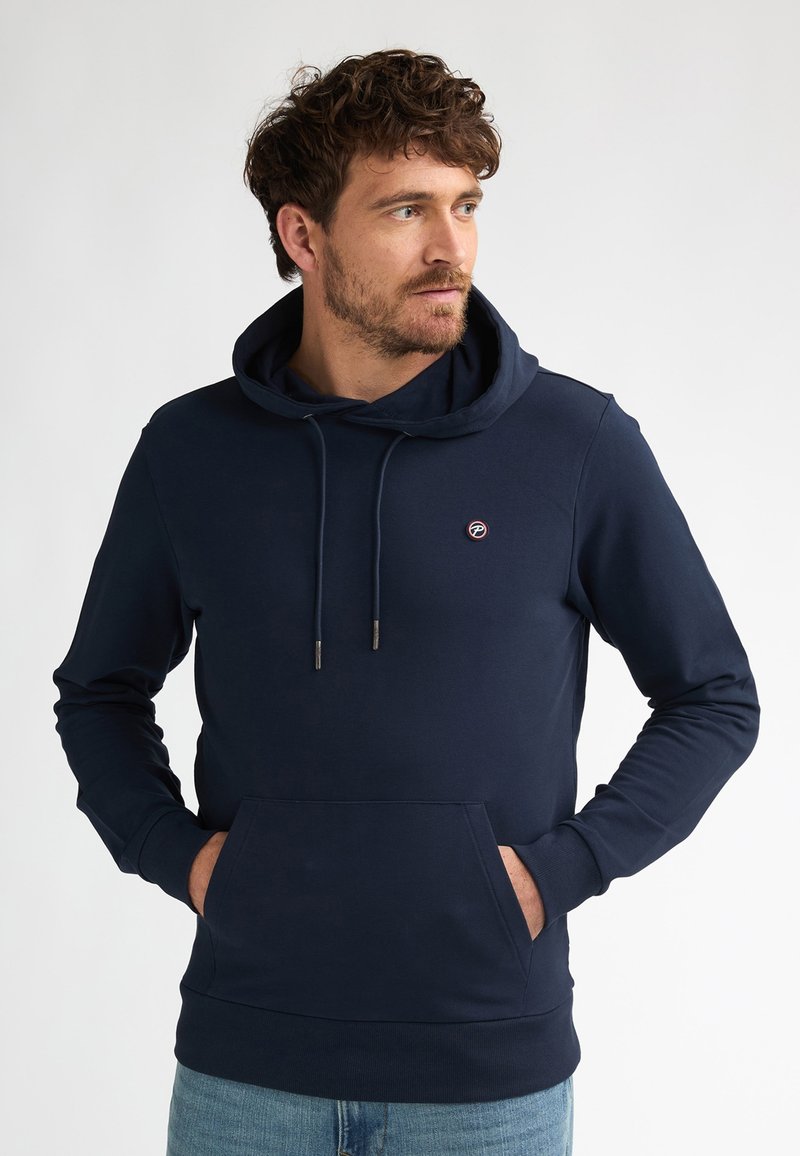 Hombre con cabello rizado que lleva una sudadera con capucha azul marino con bolsillo frontal y pequeño logo, mirando hacia la derecha con las manos en los bolsillos contra un fondo liso.