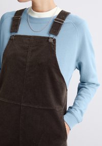 Braune Corduroy-Overalls mit verstellbaren Trägern, getragen über einem hellblauen Langarmshirt. Mit seitlichen Taschen und silberfarbenen Beschlägen.
