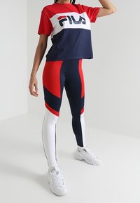 Červené a námořnické zkrácené tričko s logem Fila; odpovídající legíny s červeným, námořnickým a bílým geometrickým barevným blokováním; bílé tenisky.