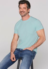 Svetlo modra kratka majica s krožnim izrezom in rahlim detajlom na rami, kombinirana s temno kavbojkami, sedi na stol. 