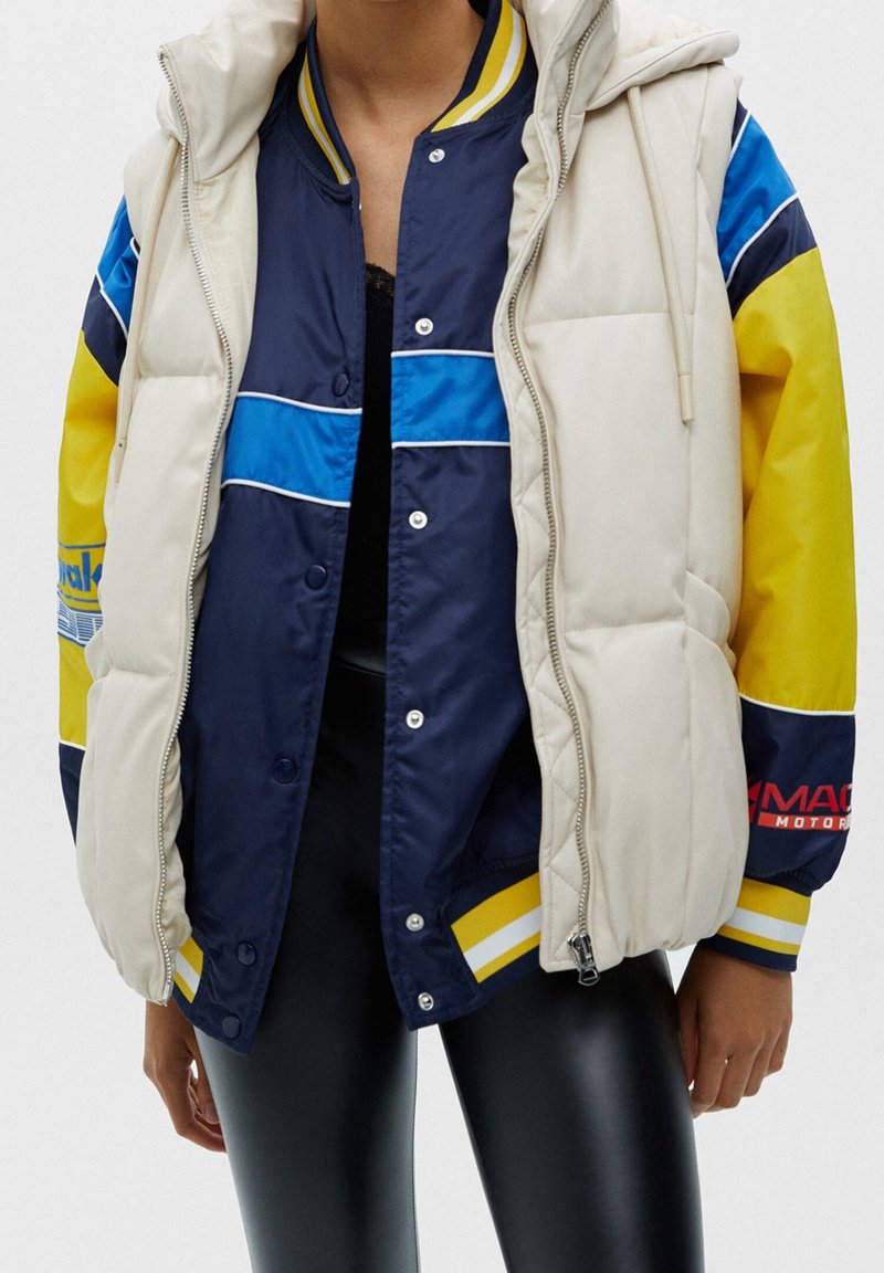 Personne portant un gilet matelassé crème par-dessus une veste universitaire à blocs de couleurs bleu, jaune et noir, et des leggings noirs brillants.