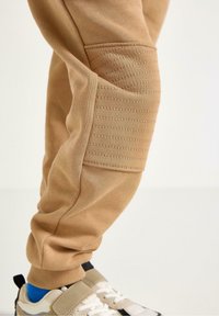 Beige Sweatpants mit gerippten Nähten an den Knien, Bündchen, und einer weichen Textur, kombiniert mit beige-blauen Sneakern.