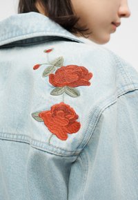 Φωτεινό γαλάζιο τζιν μπουφάν με κόκκινα κεντητά τριαντάφυλλα και πράσινα φύλλα στην πίσω πλευρά των ώμων, φορεμένο από άτομο που είναι γυρισμένο στο πλάι.