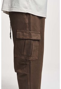 Pantalons cargo marron en tissu texturé, présentant une grande poche latérale avec un rabat et des détails en cordon de serrage à la taille.