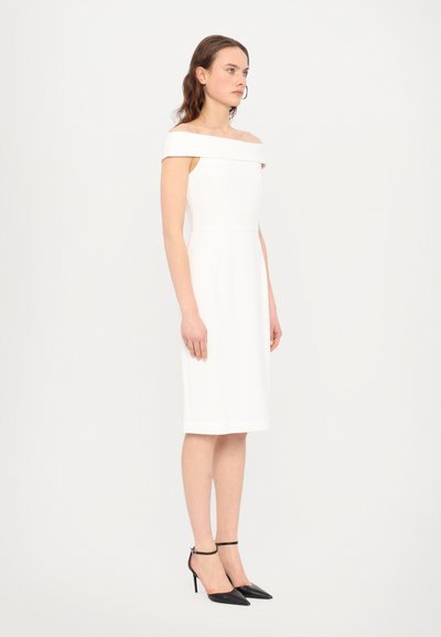 Vestido branco de ombros descobertos com uma silhueta ajustada e comprimento até ao joelho, combinado com sapatos de salto preto com bico fino e uma tira em volta do tornozelo.