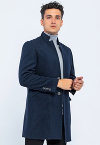 Marineblauwe wollen jas met een hoge kraag, knopen aan de voorkant, twee zijzakken en lange mouwen. G gedragen over een lichtgrijze coltrui.