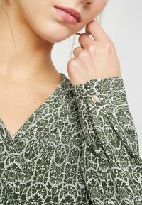 Grön mönstrad blus med vitt blommönster. V-ringning och detaljer med knapp vid ärmslutet. Mjuk tyg med en något texturerad yta.