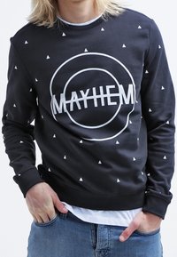 Felpa nera con motivi triangolari bianchi e una grafica centrale "MAYHEM". I polsini e l'orlo a coste completano il design casual.
