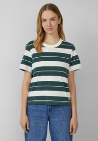 Vrouw met lichtbruin haar, gekleed in een donkergroen en wit horizontaal gestreept t-shirt en blauwe jeans, tegen een effen grijze achtergrond.