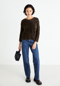 Brauner, flauschiger Strickpullover mit V-Ausschnitt, getragen mit blauen Jeans im geraden Schnitt und schwarzen, klobigen Plateaustiefeln. Kleine schwarze Handtasche in der Hand.