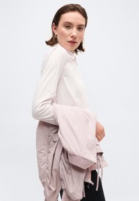 Νεαρή γυναίκα με κοντά καστανά μαλλιά φορώντας λευκό πουκάμισο και κρατώντας ένα ανοιχτό ροζ σακάκι στον βραχίονα της, μπροστά σε απλό φόντο.