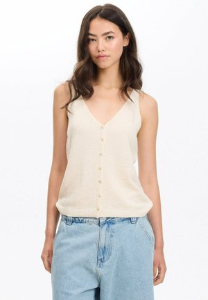 Joven con cabello largo y oscuro que lleva una blusa sin mangas cremosa con botones y vaqueros de talle alto azul claro, de pie frente a un fondo blanco.