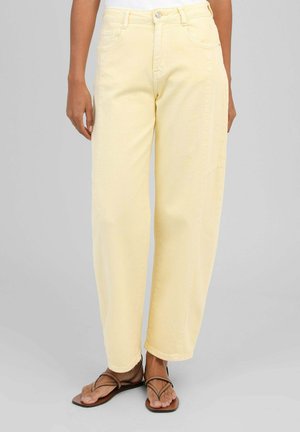 Personne portant un pantalon jaune pâle taille haute, coupe droite, et des sandales marron à lanières, debout devant un fond gris uni.