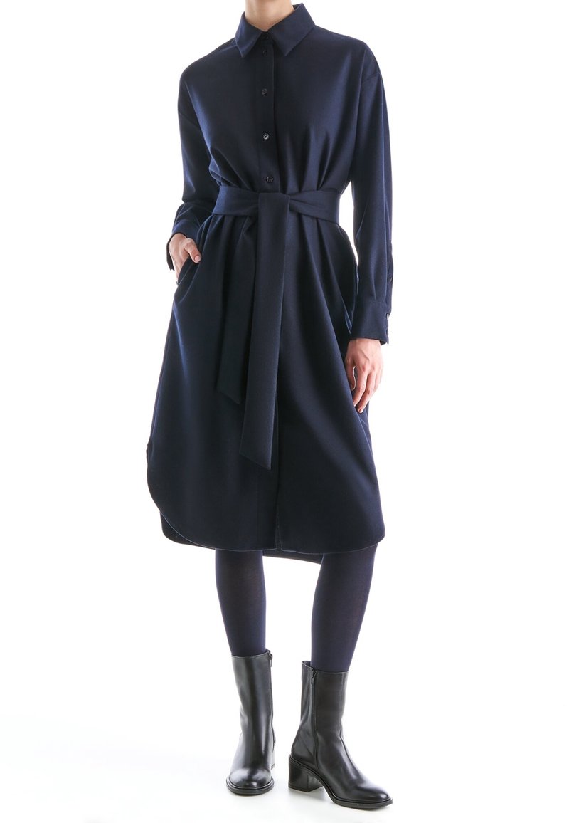 Femme portant une robe chemise ceinturée à manches longues bleu marine avec des poches, associée à des collants bleu marine et des bottines noires.