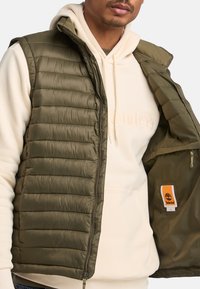 Olijfgroen gewatteerd vest met een ritssluiting, voorzien van een verticale quiltstructuur. Gewatteerde binnenvoering met een zichtbaar Timberland logo label.