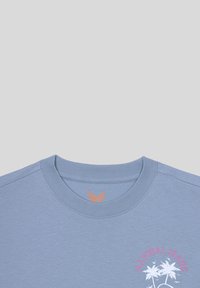 T-shirt bleu clair à manches courtes en coton. Il présente un col ras du cou et un imprimé de palmiers avec le texte "Kaporal Jeans" en rose.