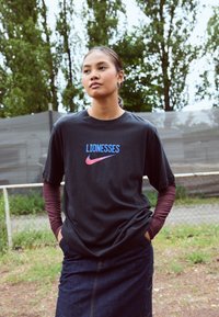 Camiseta de algodón negra con "LIONESSES" y logo de Nike en degradado de azul a rosa. Llevada sobre una camisa de manga larga burdeos. Falda de denim abajo.