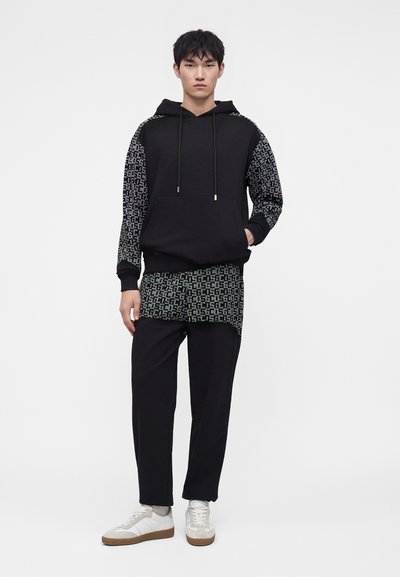 Jeune homme debout portant un sweat à capuche noir avec des manches et un ourlet à motifs, un pantalon noir et des baskets blanches sur un fond blanc uni.