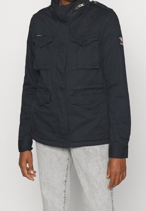 Personne portant une veste noire avec quatre poches à rabat à l'avant et un jean gris clair, debout devant un fond blanc uni.