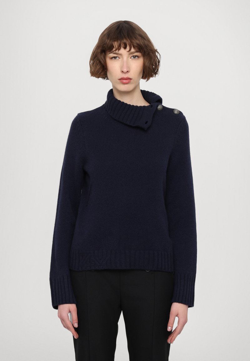 Pull en maille bleu marine avec un col montant à côtes et des boutons décoratifs sur le côté. Présente une texture unie et une coupe légèrement courte avec des poignets côtelés.