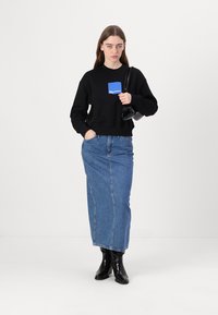 Felpa corta nera con logo blu, abbinata a lunga gonna di jeans blu e stivali neri alla caviglia. Il modello tiene in mano una piccola borsa nera.