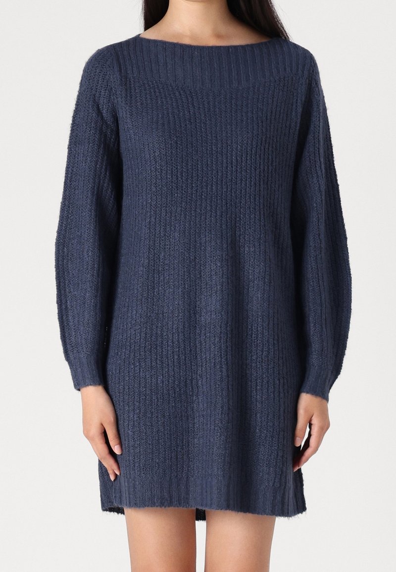 Femme portant une robe-pull tricotée bleu marine à manches longues avec un col et des poignets côtelés, debout devant un fond uni.