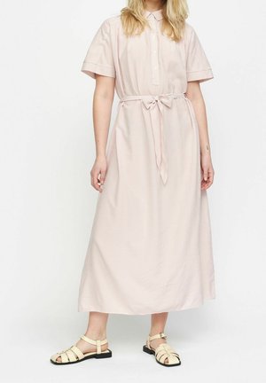 Femme portant une robe midi rose clair à manches courtes avec une ceinture nouée devant et des sandales plates beiges à fines lanières sur fond blanc.
