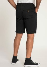 Shorts noirs avec bords roulés, en tissu de coton. Présentent deux poches arrière avec fermetures à bouton et une coupe décontractée. Portés avec des baskets montantes noires.