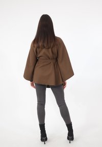 Indumento esterno marrone con cintura e maniche a campana, sovrapposto a pantaloni grigi aderenti e stivali neri con tacco, visto da dietro su sfondo bianco.