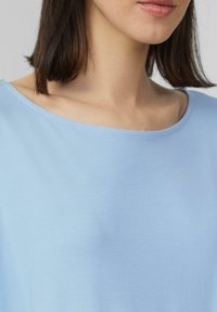 Lichtblauwe blouse met een brede, ronde halslijn. Zacht materiaal, gladde textuur en een casual pasvorm. Geen zichtbare patronen of hardware details.