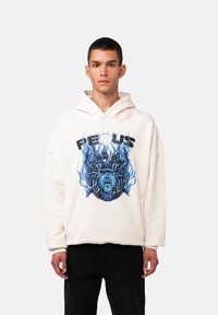 Felpa crema con un design grafico che presenta un volto illustrato blu e nero circondato da fiamme. Materiale in cotone, vestibilità rilassata e tasca a marsupio.
