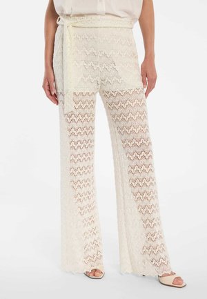 Femme portant un pantalon large en dentelle blanche avec une ceinture assortie, associé à des sandales beiges à bout ouvert sur un fond blanc.