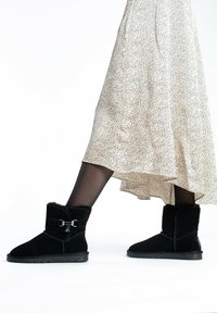 Botines de ante negro con forro de felpa, que cuentan con un lazo decorativo y acentos de metal plateado. Se llevan con una falda fluida y estampada.
