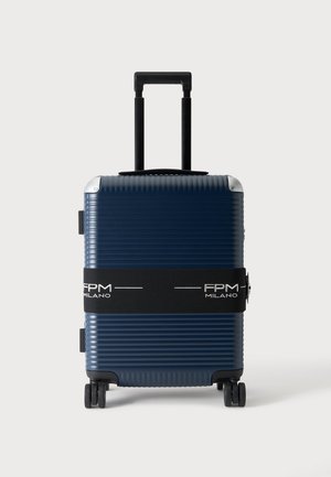 Valise coque rigide bleu marine avec des rainures horizontales, poignée escamotable noire, quatre roues noires et une sangle noire marquée FPM Milano.