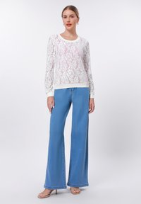 Λευκή δαντέλα μπλούζα με μακριά μανίκια και λουλουδάτο σχέδιο, συνδυασμένη με ανοιχτό μπλε καμπάνα τζιν. Το outfit έχει στρογγυλή λαιμόκοψη και χαλαρή εφαρμογή.