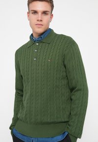Pull en maille torsadée vert avec un col et une patte de boutonnage à trois boutons, porté avec une chemise en denim bleue en dessous.