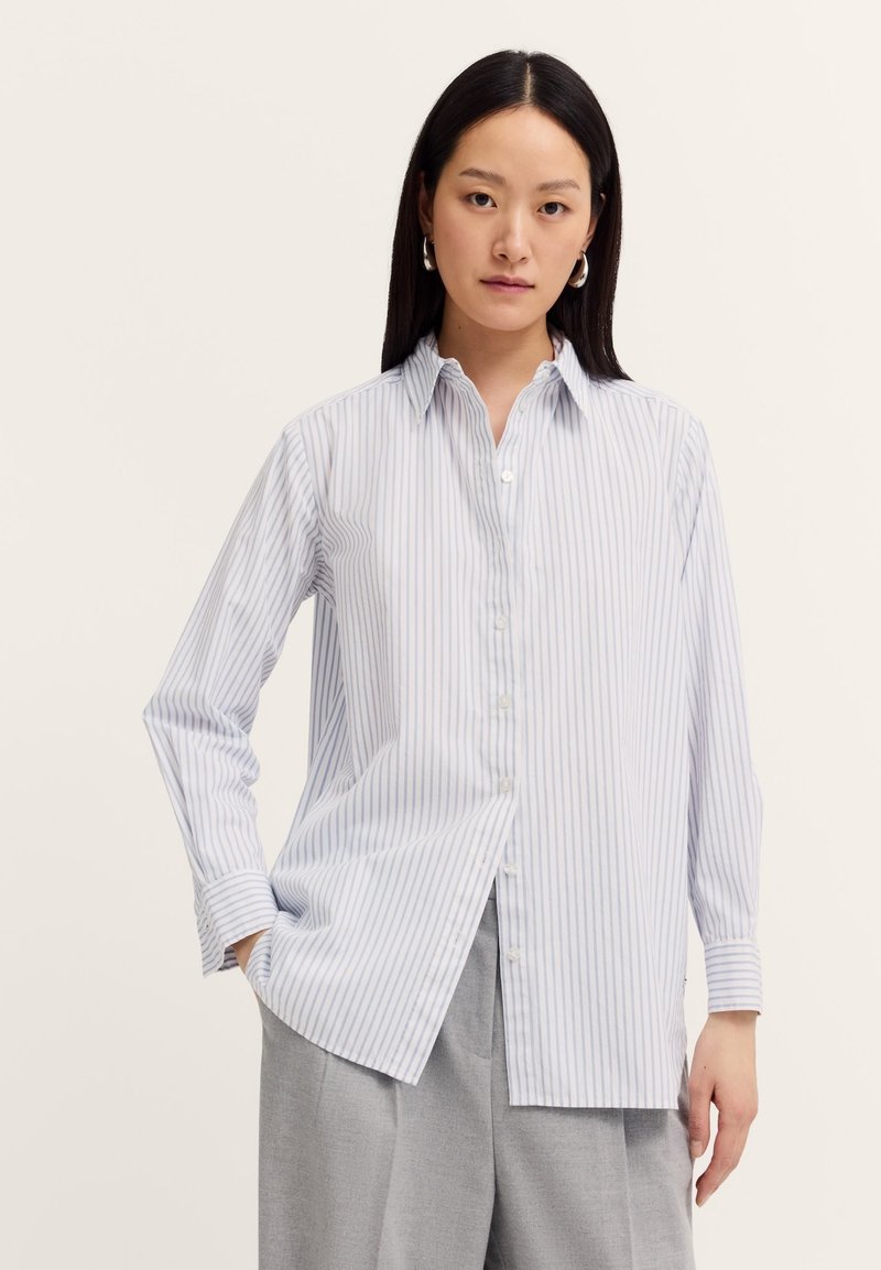 A woman stands confidently, wearing a light blue and white striped shirt, with gray pants. She has straight black hair and simple earrings.